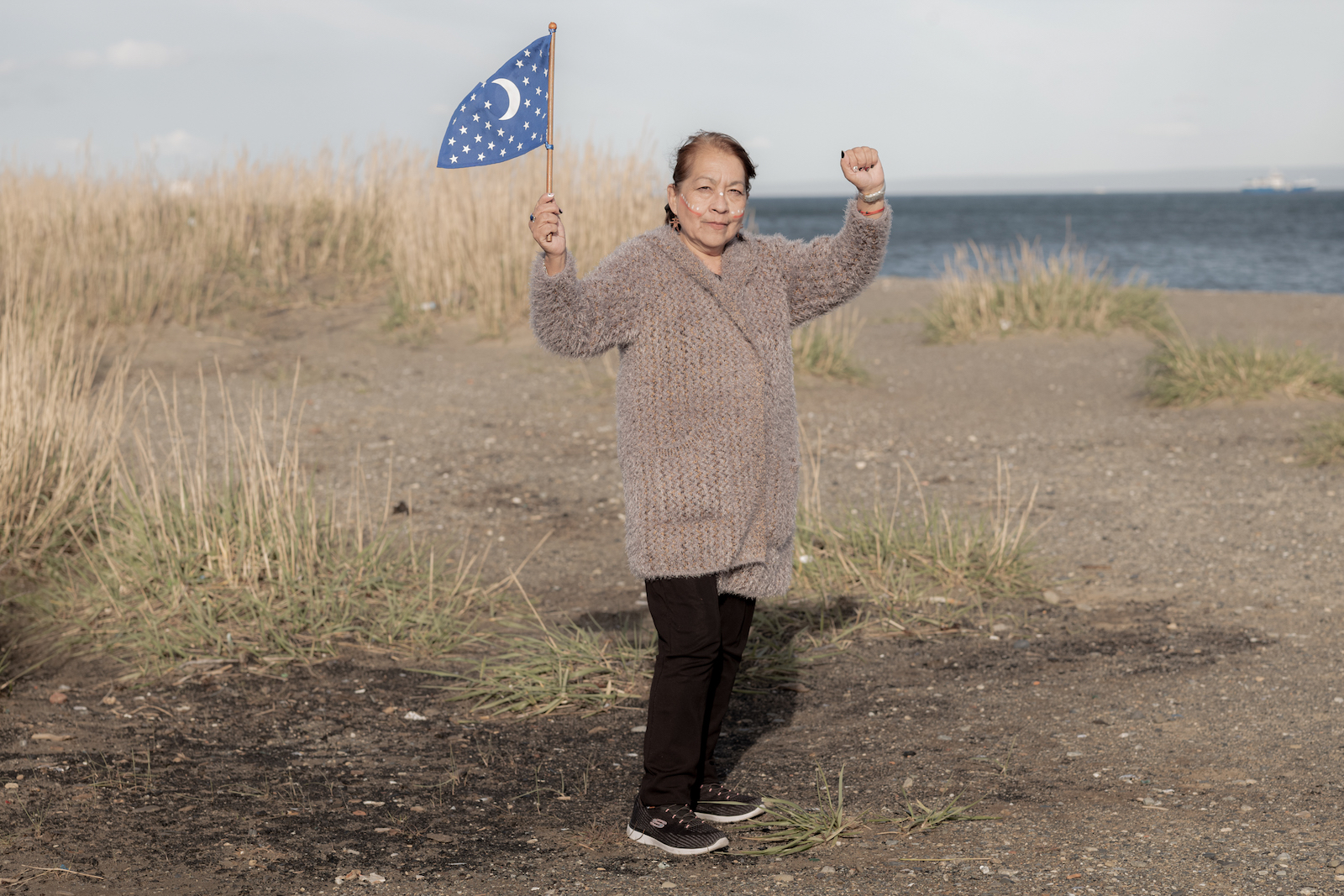 A orillas del Estrecho de Magallanes, María Margarita Vásquez Choque (Pilar) sostiene con orgullo la bandera que representa a los Selk'nam y levanta el puño en señal de lucha y resistencia. Punta Arenas, Chile, 2021. Foto: Marcio Pimenta